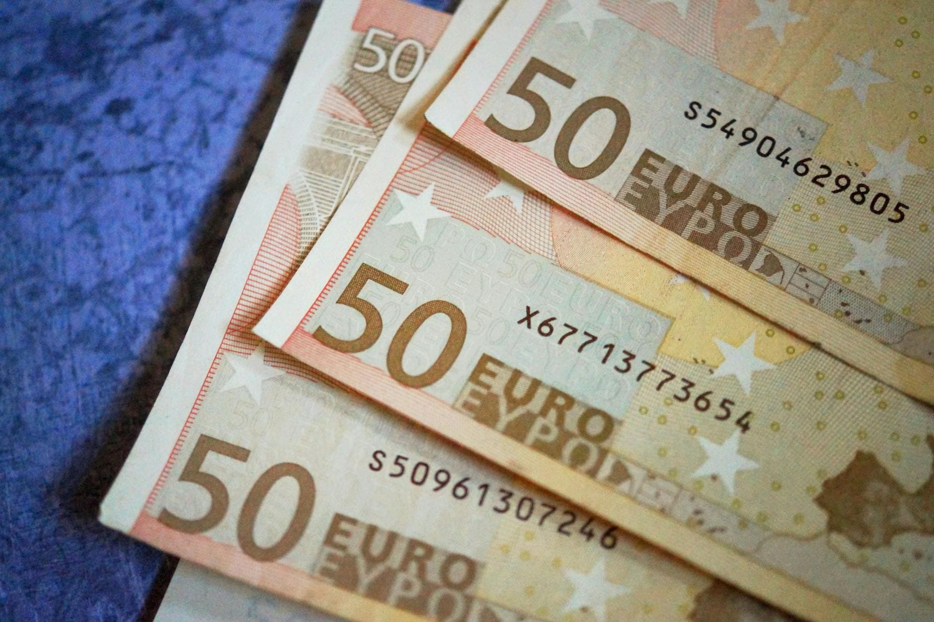 Mehrere gefächerte 50-Euro-Banknoten mit sichtbaren Seriennummern liegen auf einem blauen, strukturierten Untergrund.