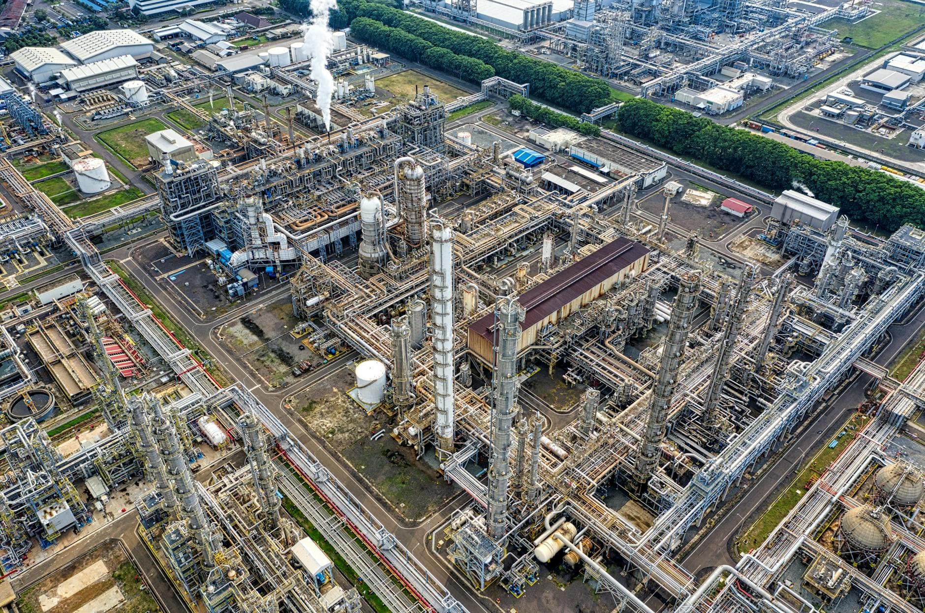 Luftaufnahme einer weitläufigen Industrieanlage mit vielen hohen Türmen, Rohrleitungen und Gebäuden, Rauch steigt auf.