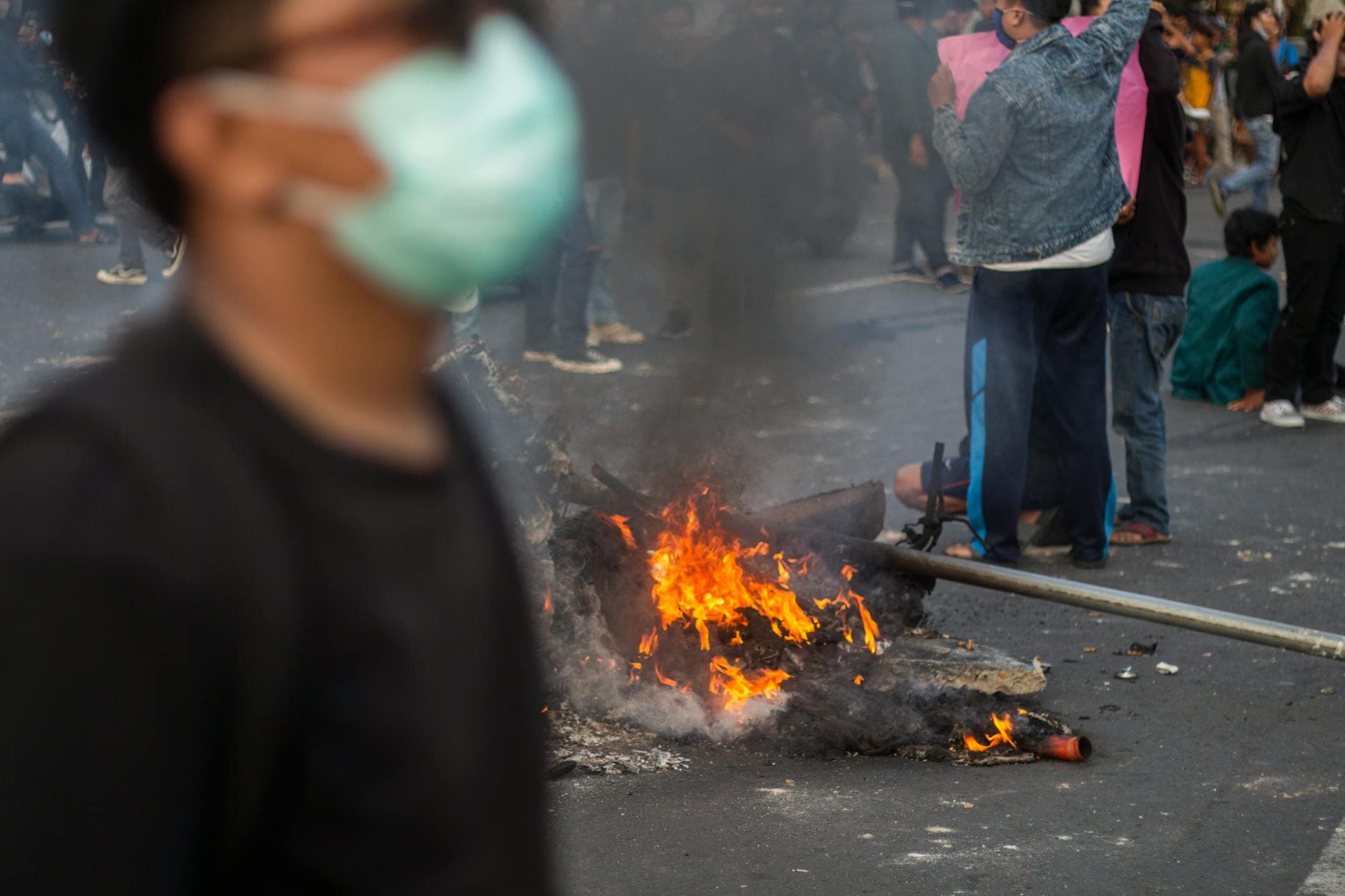 Unscharf im Vordergrund: Person mit blauer Maske; Mitte: Feuer auf Asphalt mit Rauch; Hintergrund: Protestierende, einige mit rosa Plakaten.