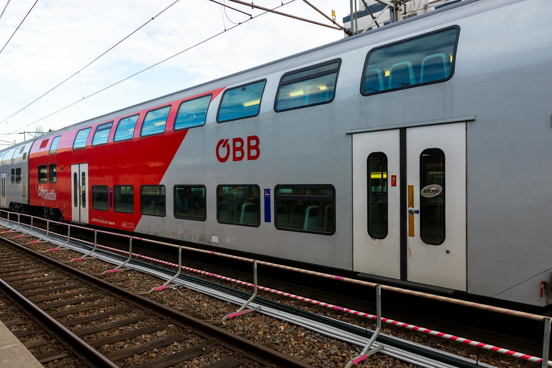 Roter und silberner ÖBB-Doppelstockzug "CityShuttle" an einem Gleis mit Oberleitung und Absperrung.