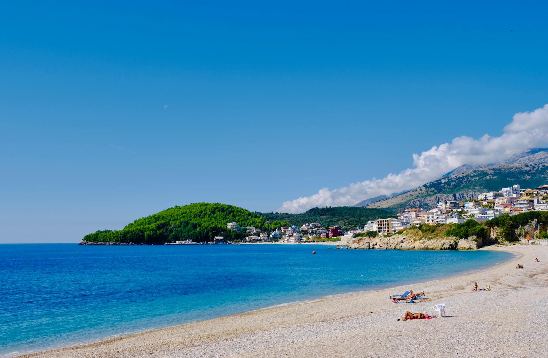 Heller Strand mit Badegästen am blauen Meer, einer Küstenstadt, grünen Hügeln und Bergen unter blauem Himmel mit Mond.