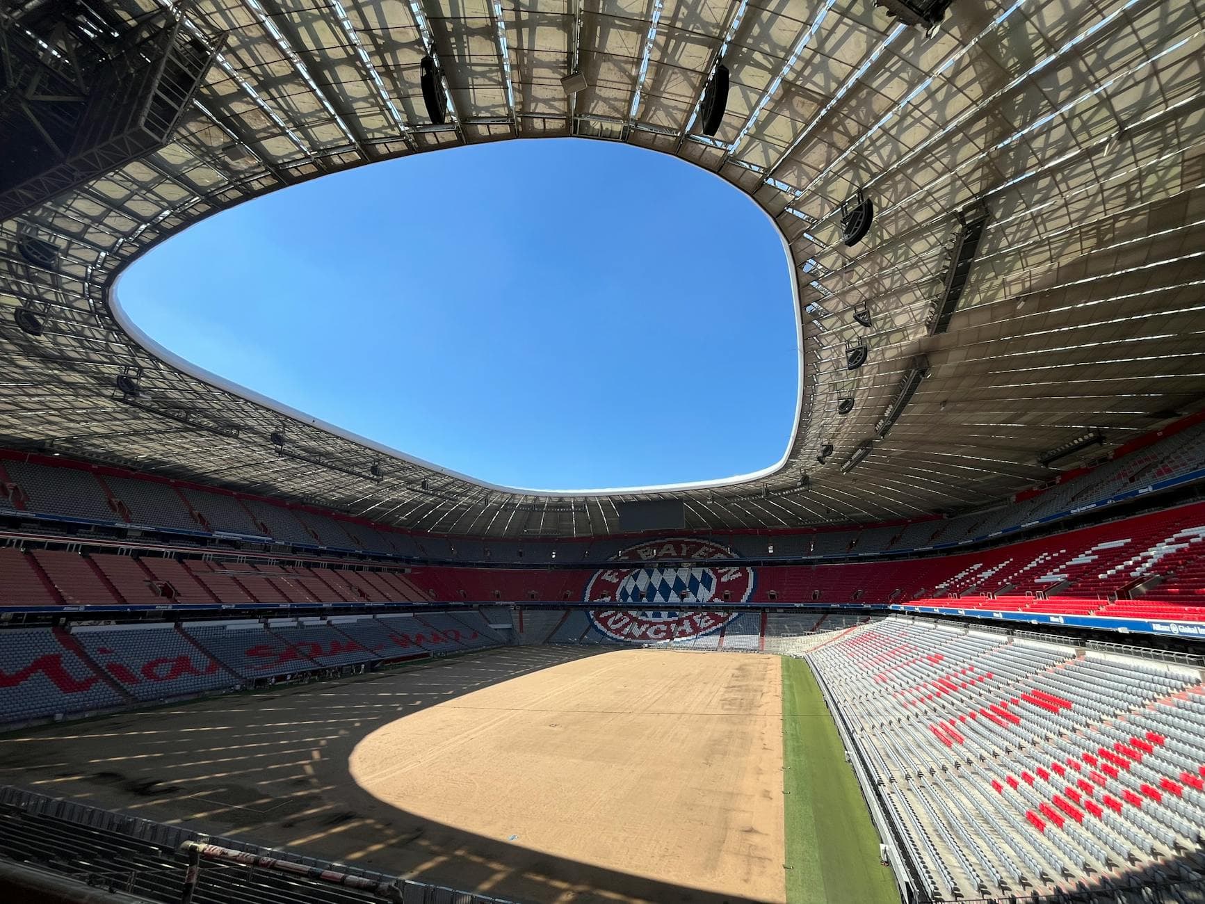 Leeres Fußballstadion bei blauem Himmel, mit sandigem Spielfeld, offenem Dach und roten und grauen Tribünen samt Bayern-Logo.