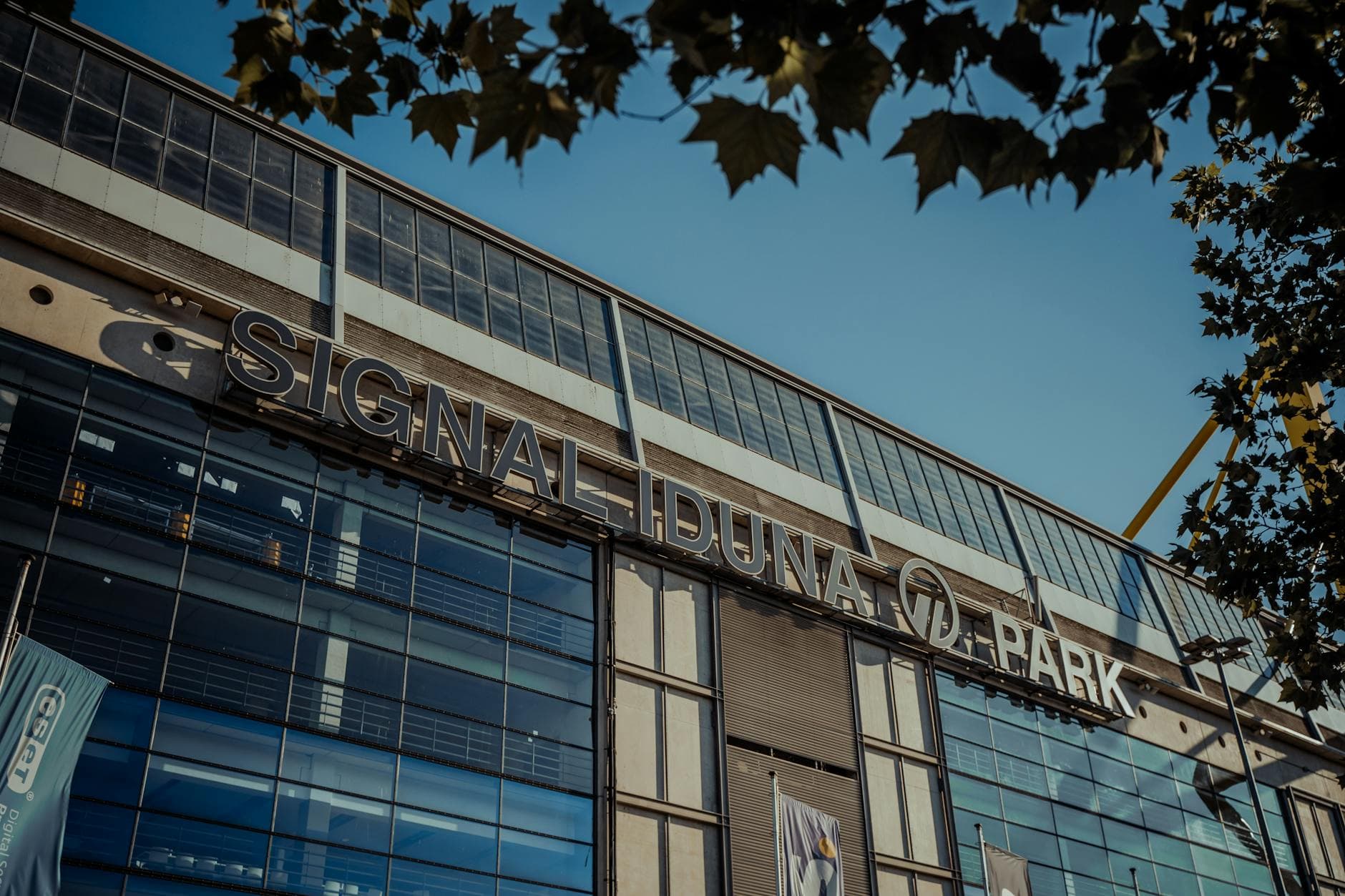 Signal Iduna Park Fassade mit Schriftzug unter blauem Himmel, teilweise von Laub verdeckt, gelbe Stadionstruktur sichtbar.