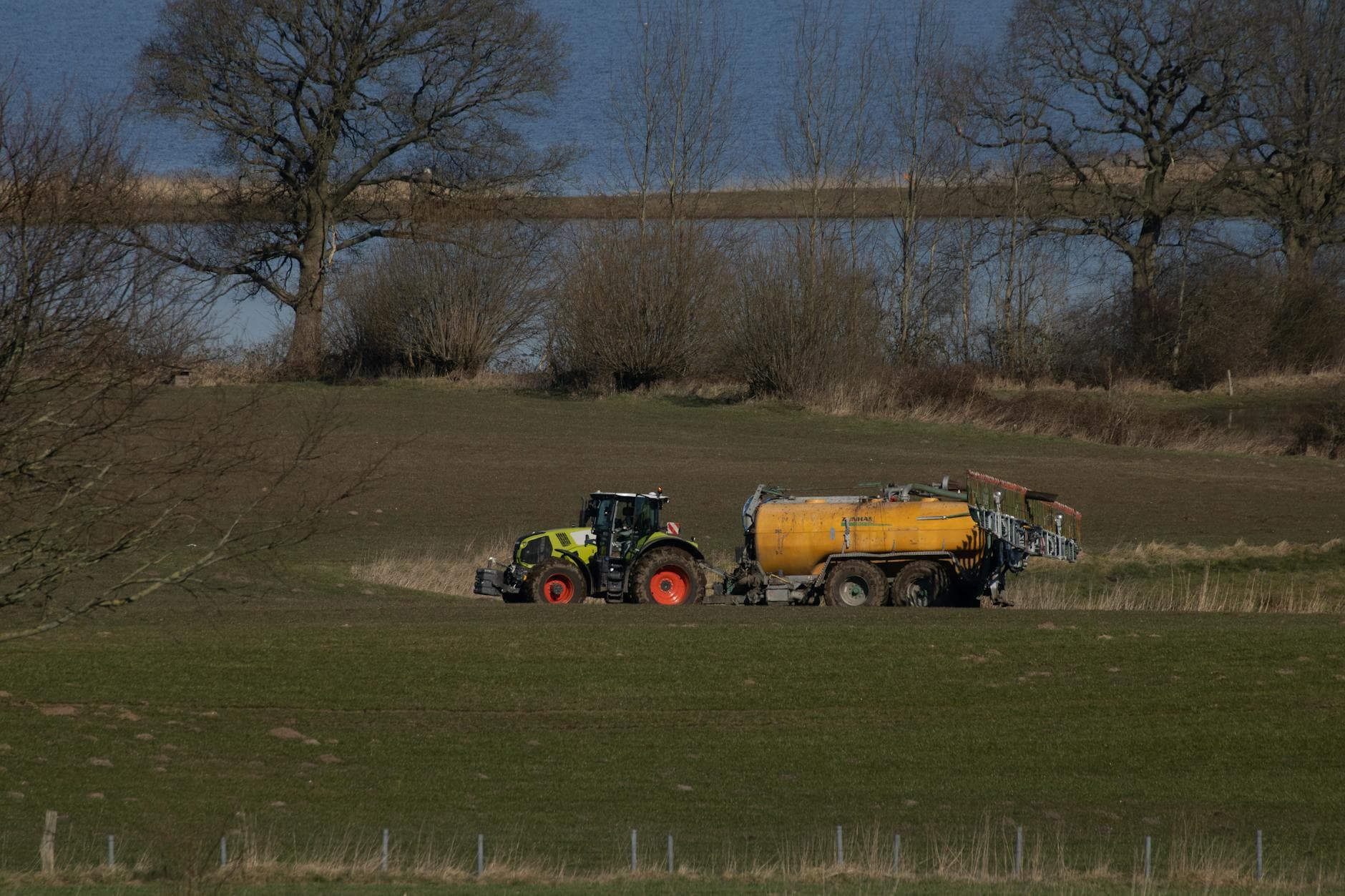 Grüner Traktor mit gelbem Güllefass auf einem Feld vor einem See mit kahlen Bäumen am Ufer.