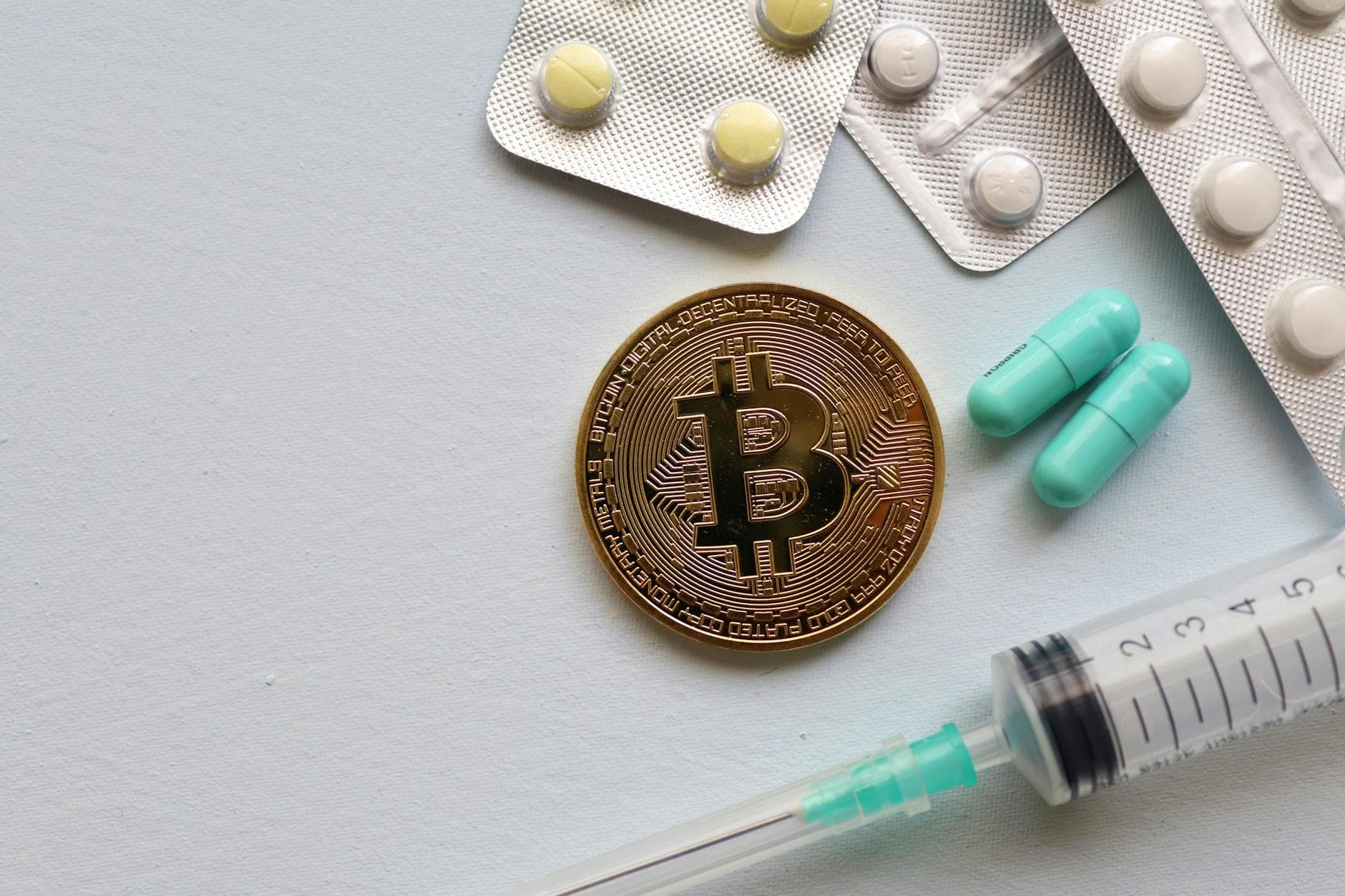 Ein goldener Bitcoin-Coin, Tabletten in Blistern, zwei türkise Kapseln und eine Spritze auf hellem Hintergrund.