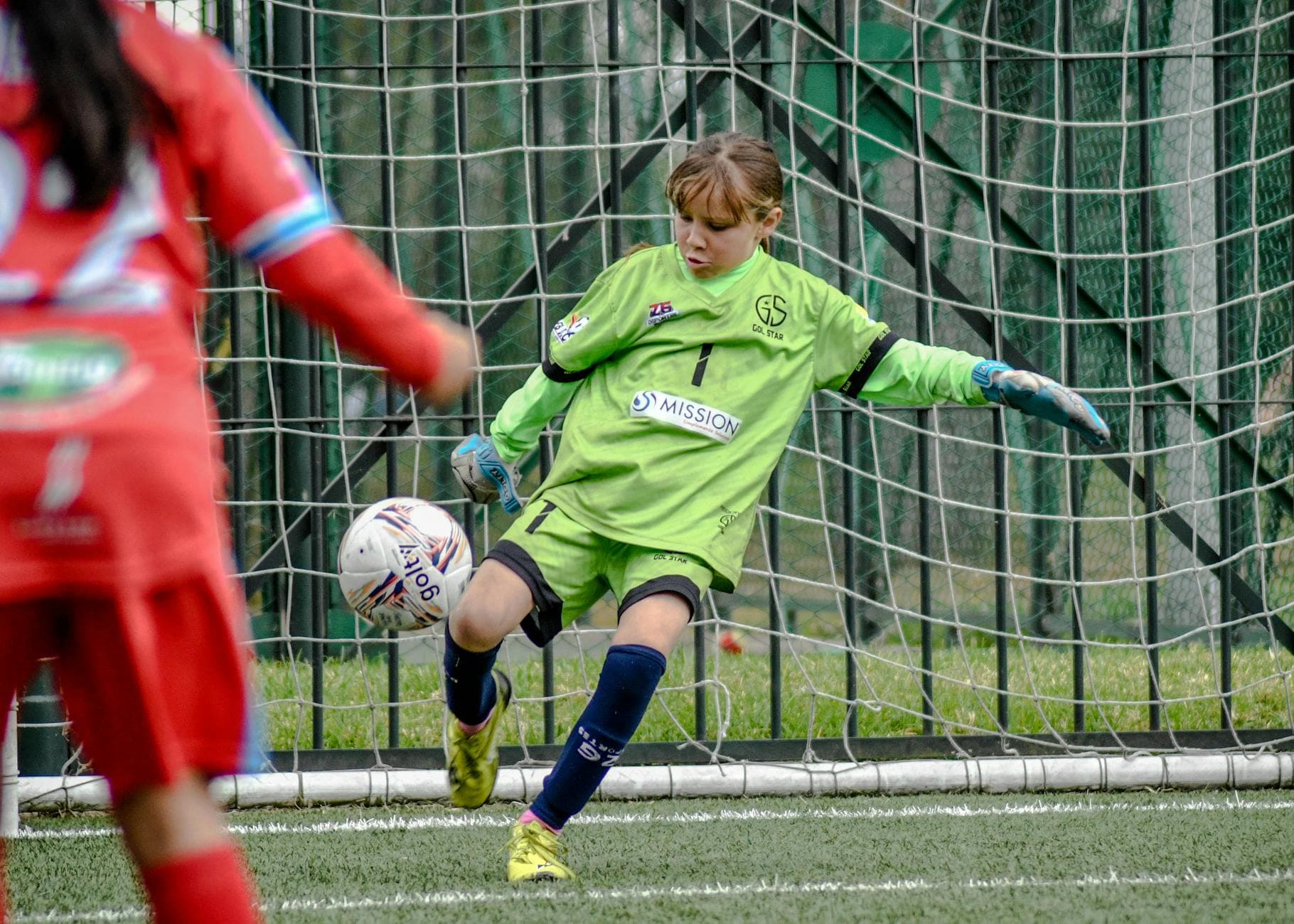 Ein junges Mädchen als Fußballtorhüterin in grünem Trikot tritt einen Ball vor einem Tor; links ein rot gekleideter Spieler.