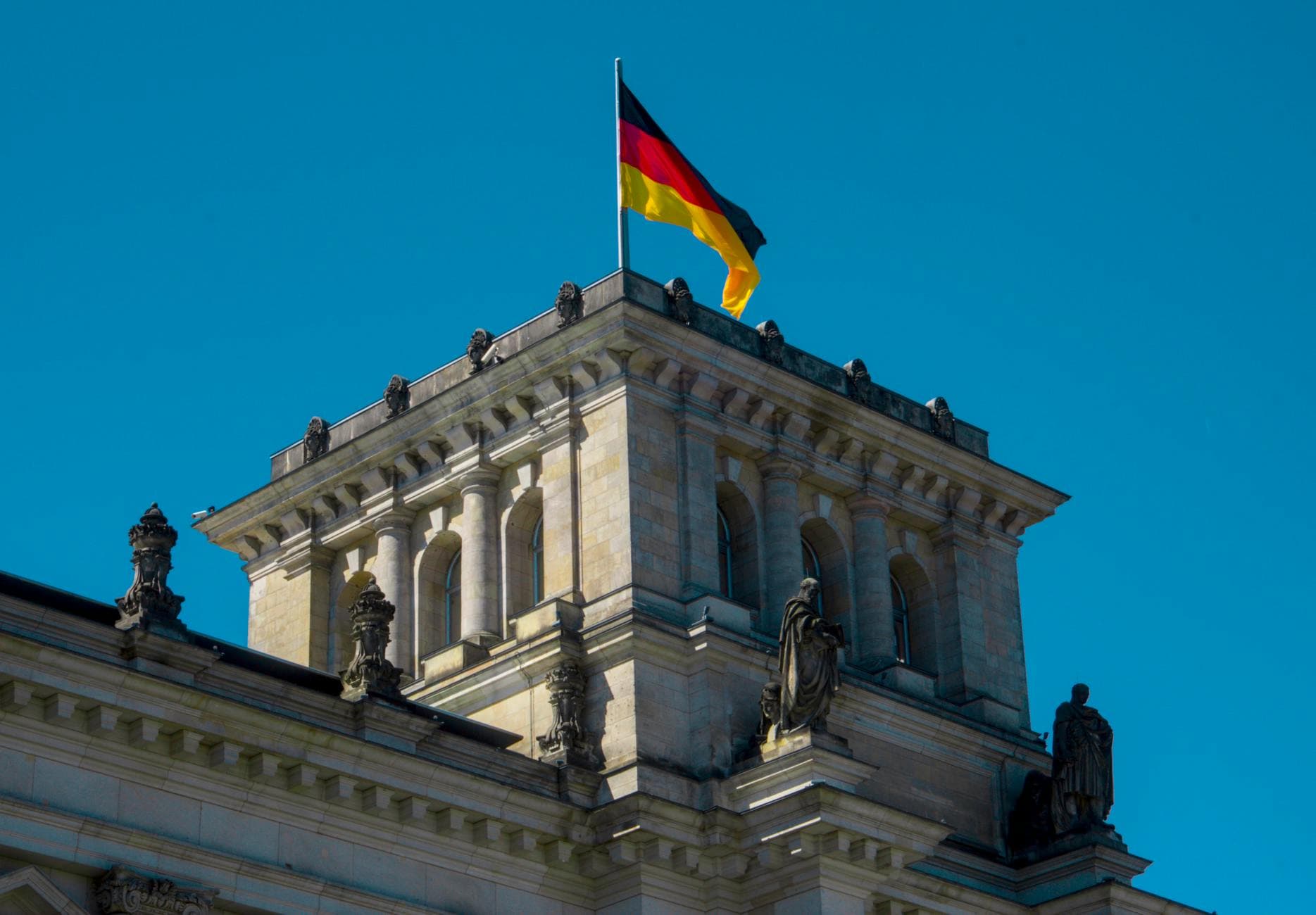 Eine wehende Deutschlandflagge auf einem monumentalen Steingebäude mit Säulen und Statuen unter blauem Himmel.