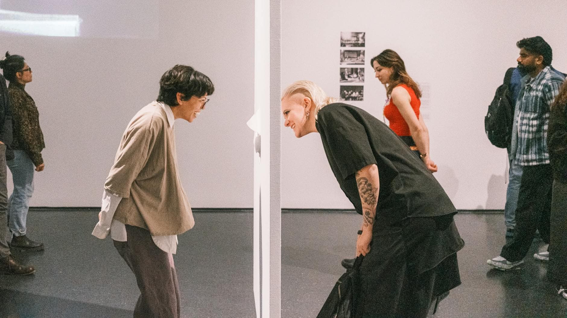 Zwei Personen beugen sich lächelnd zu einer weißen Trennwand, im Hintergrund weitere Besucher einer Ausstellung.