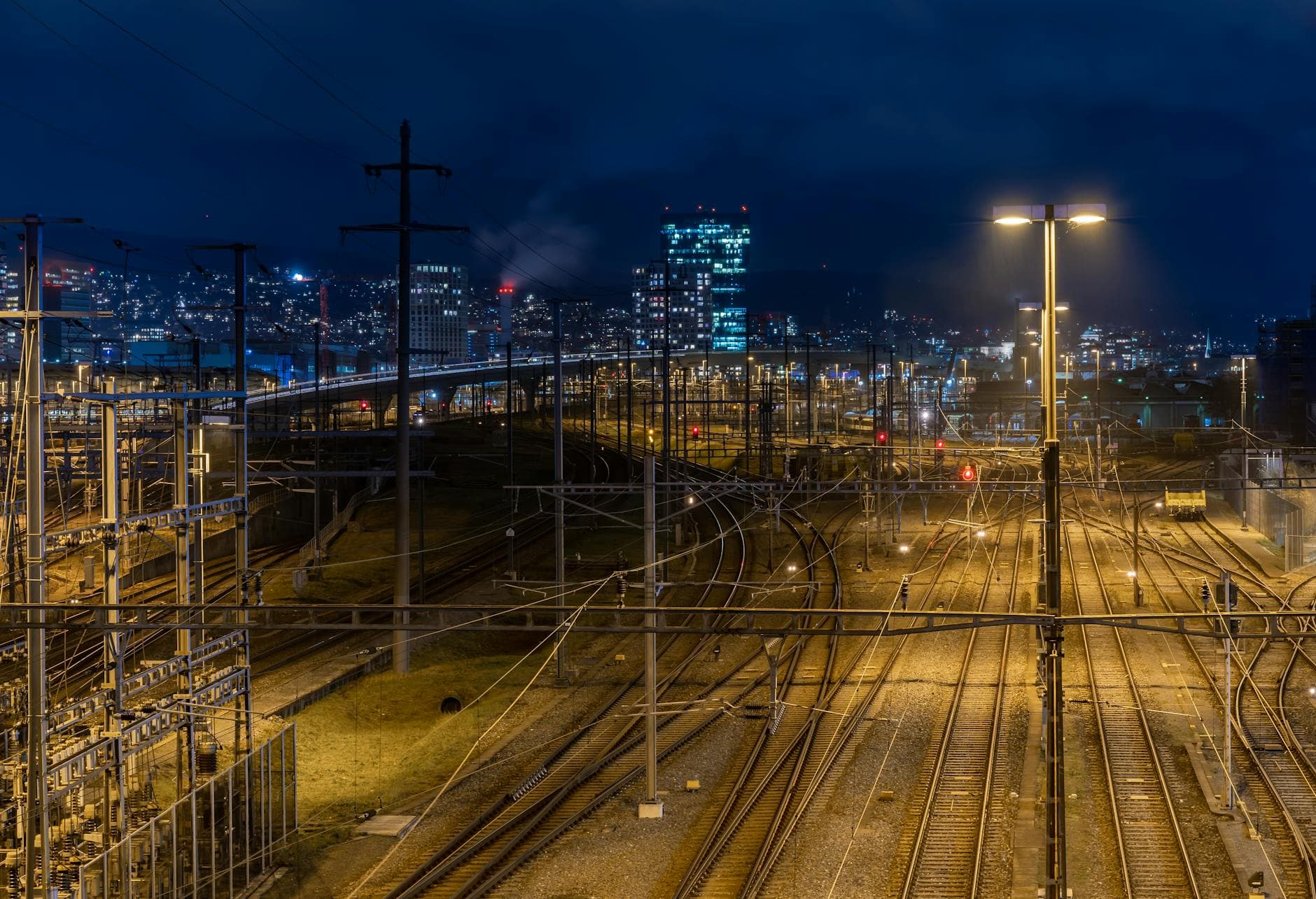 Nachtaufnahme eines beleuchteten Eisenbahnknotenpunkts mit vielen Gleisen, Oberleitungen, roten Signalen und Stadt im Hintergrund.