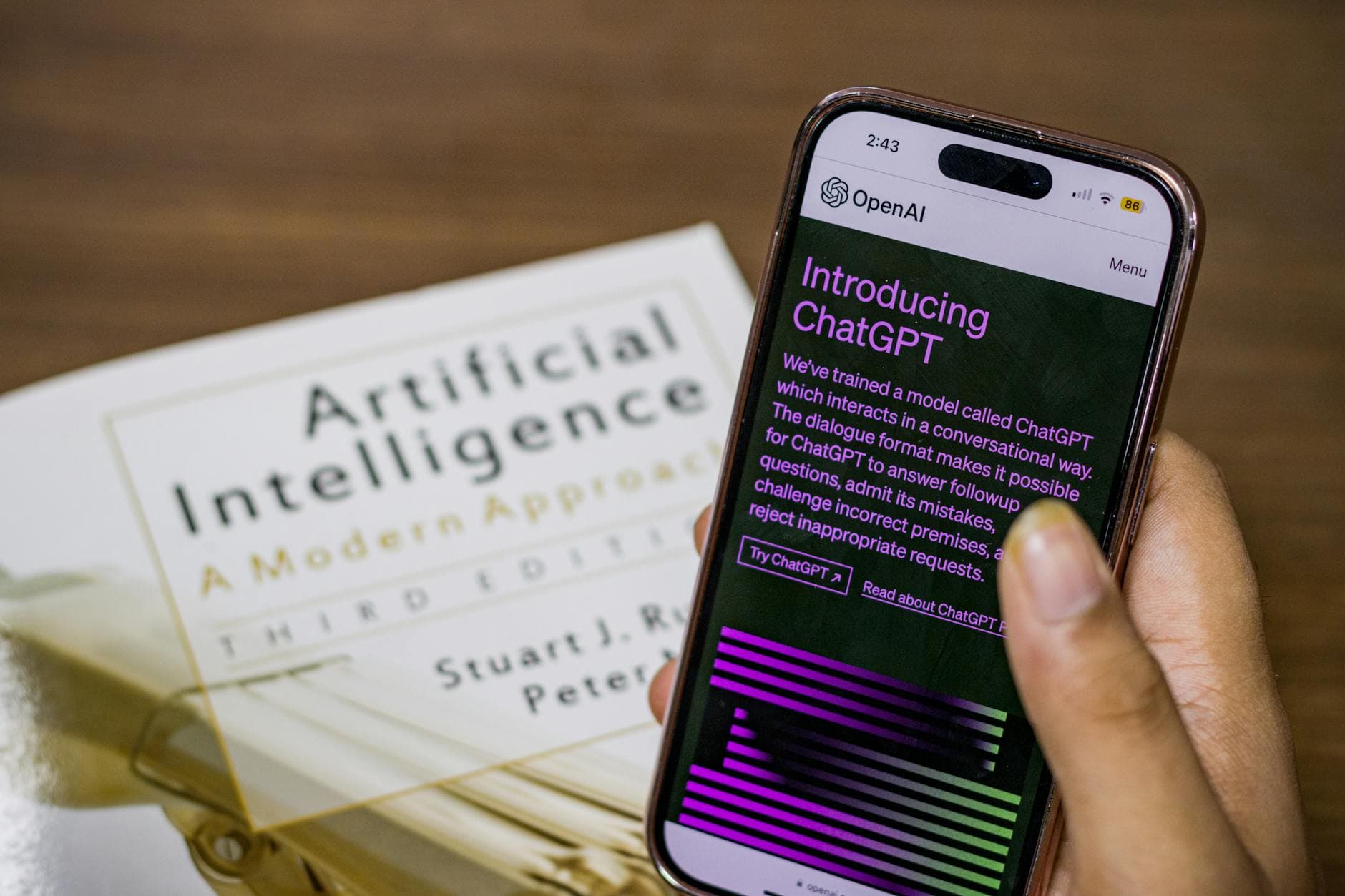 Hand hält Smartphone mit ChatGPT-Ankündigung von OpenAI, im Hintergrund ist ein Buch über Künstliche Intelligenz zu sehen.