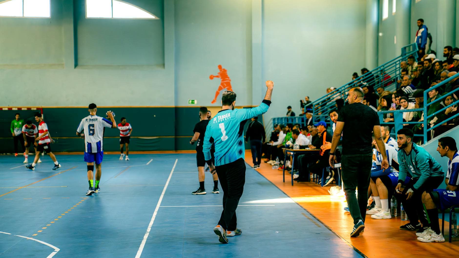 Handballspiel in einer Halle: Spieler auf blauem Feld, jubelnder Torwart, Zuschauer auf Tribünen, orange Silhouette an der Wand.