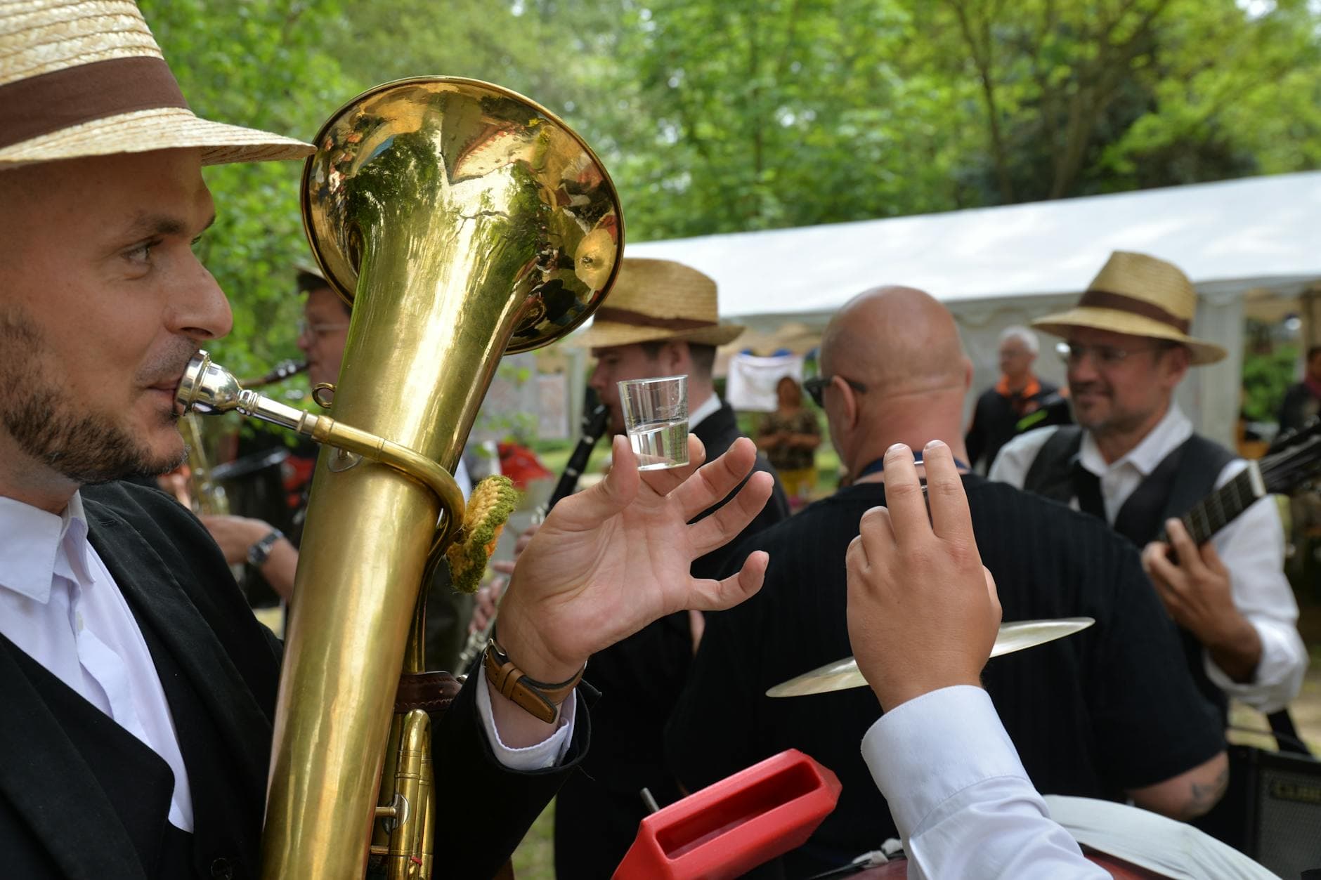 Musiker mit Strohhut spielt Tuba, hält Schnapsglas; im Vordergrund ein Becken, hinten weitere Musiker und ein Zelt.
