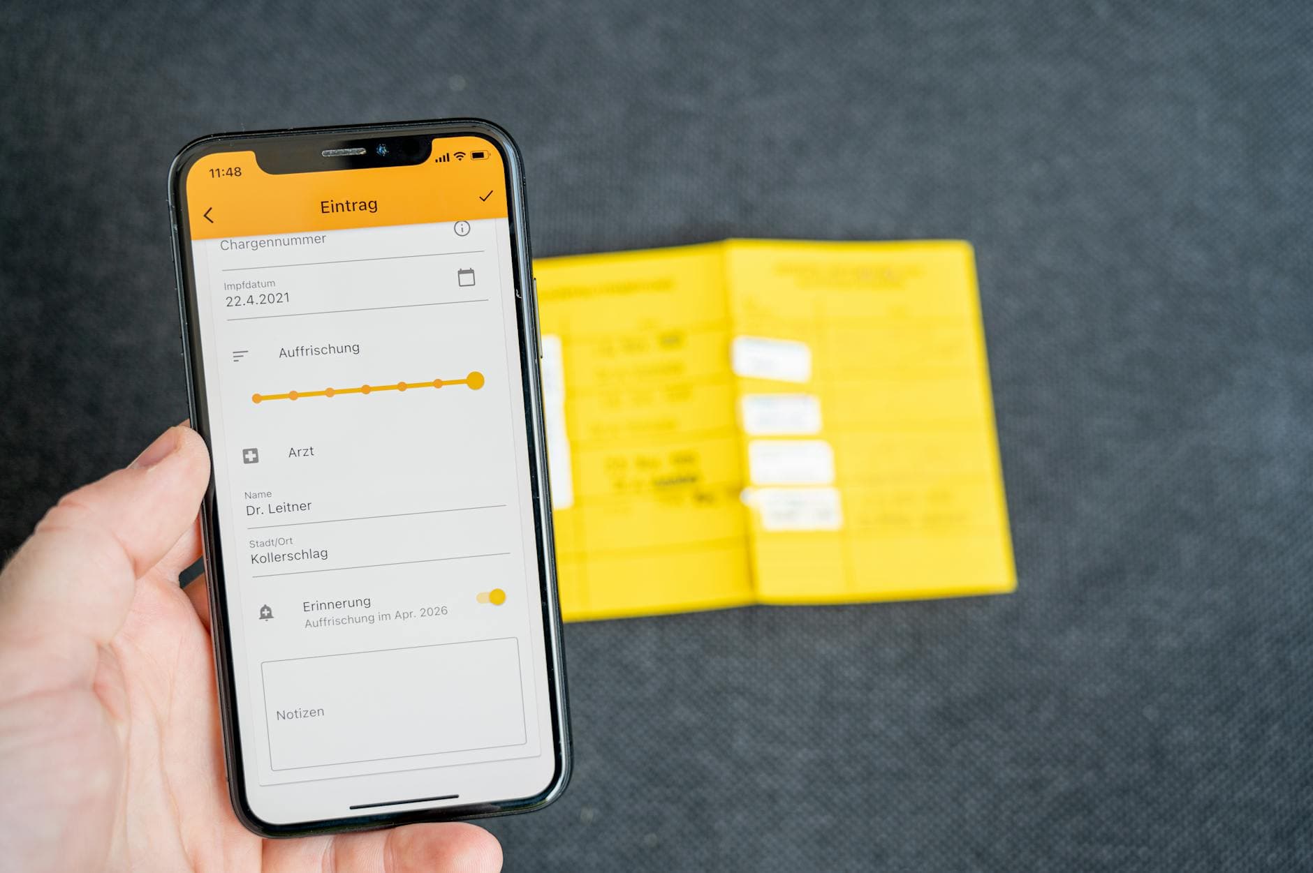 Eine Hand hält ein Smartphone mit einer Impfpass-App, die einen Eintrag vom 22.4.2021 und eine Auffrischung zeigt; im Hintergrund liegt ein gelber Impfpass.
