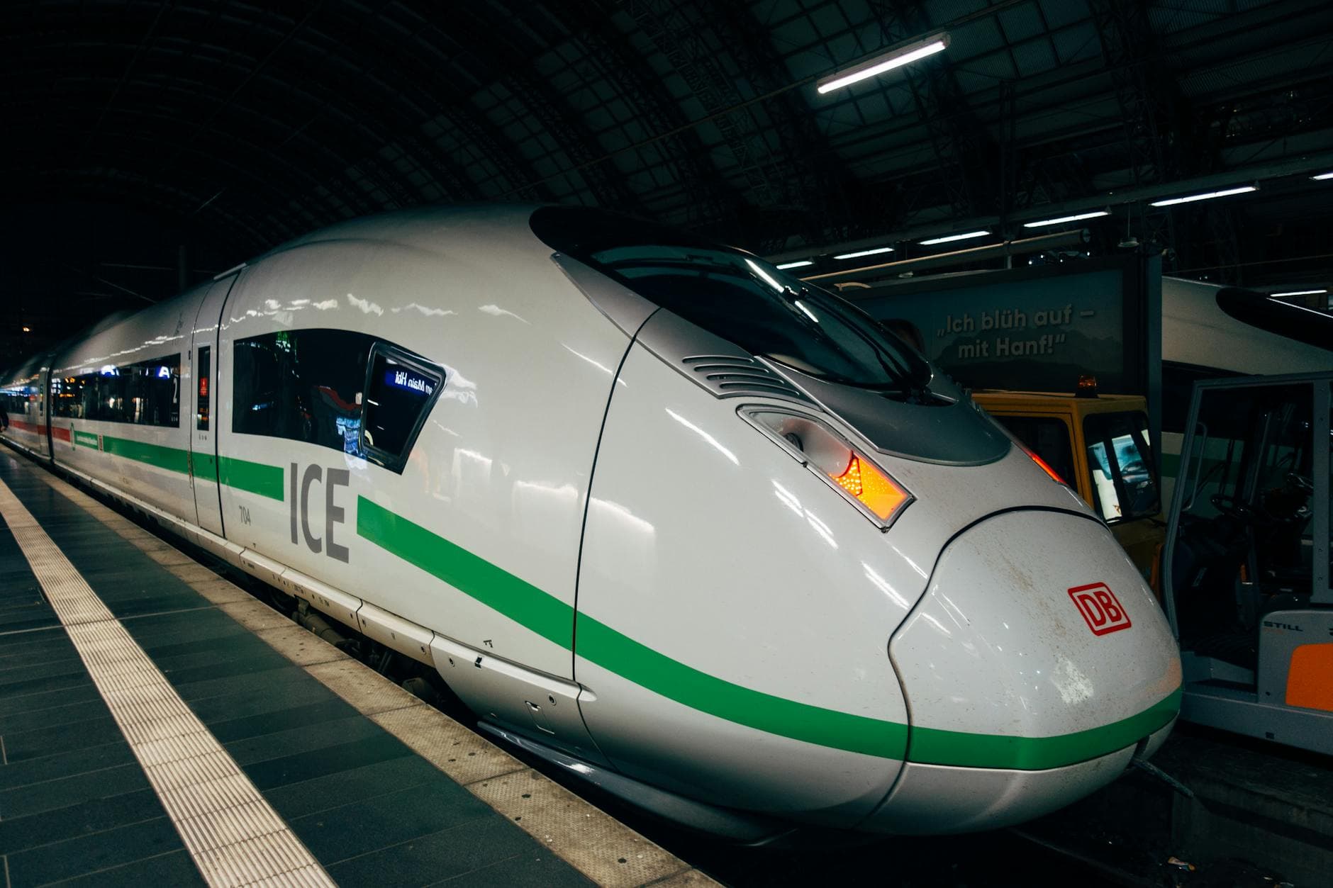 Ein weiß-grüner ICE-Zug der Deutschen Bahn steht an einem Bahnsteig; im Hintergrund ein gelbes Fahrzeug und ein Werbeschild.
