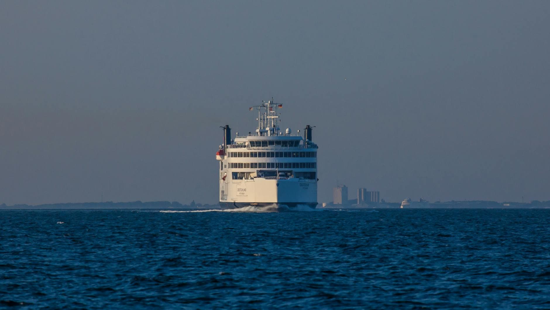 Das weiße Passagierschiff DEUTSCHLAND fährt auf blauem Meer, im Hintergrund Küstenlinie mit Stadt und einem weiteren Schiff.