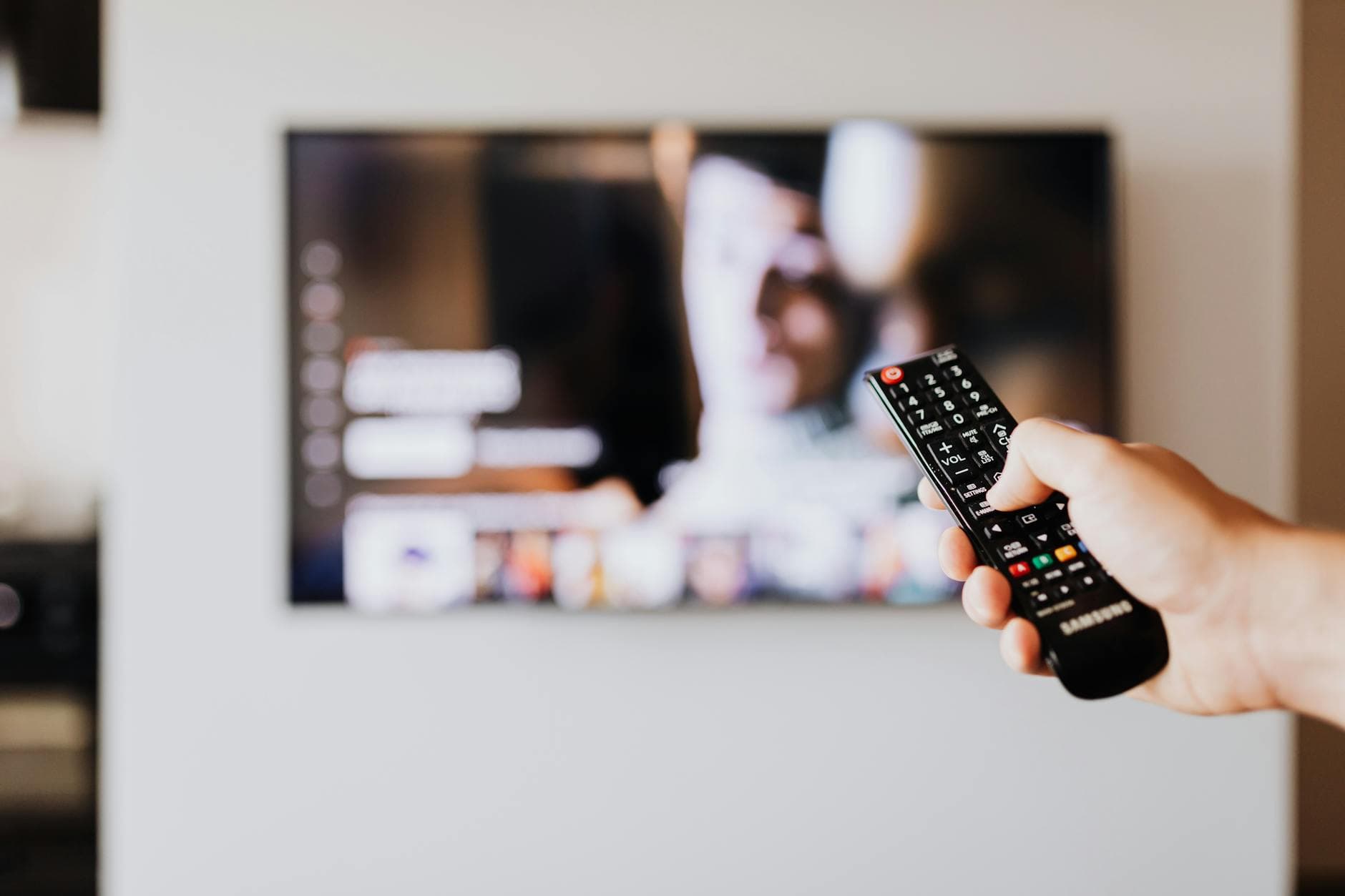 Eine Hand hält eine schwarze Samsung-Fernbedienung und drückt einen Knopf; im Hintergrund ein unscharfer TV-Bildschirm.