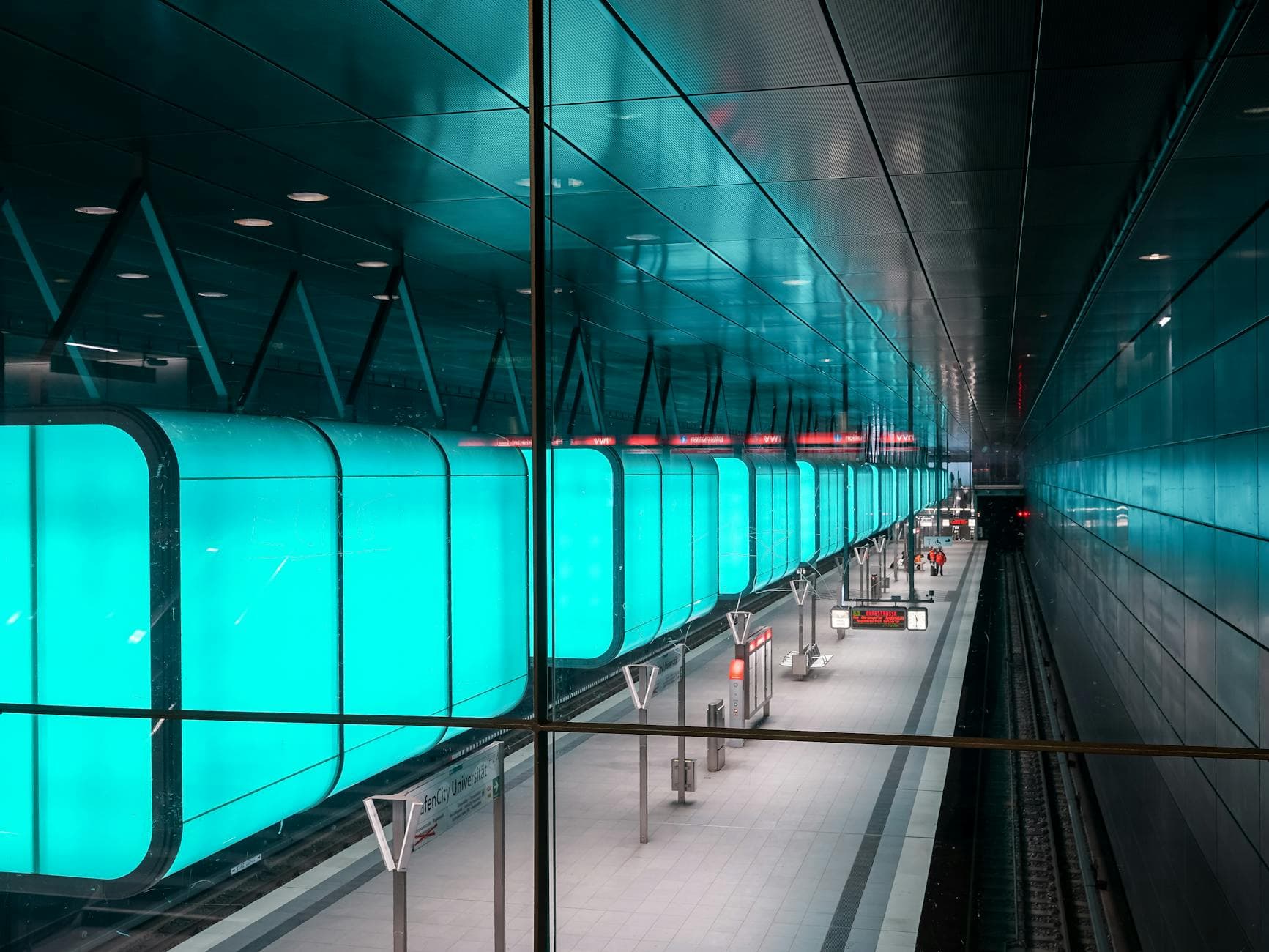 Moderner U-Bahnhof mit einem türkis leuchtenden Zug hinter Glas, Bahnsteig, Gleisen und einigen Personen.