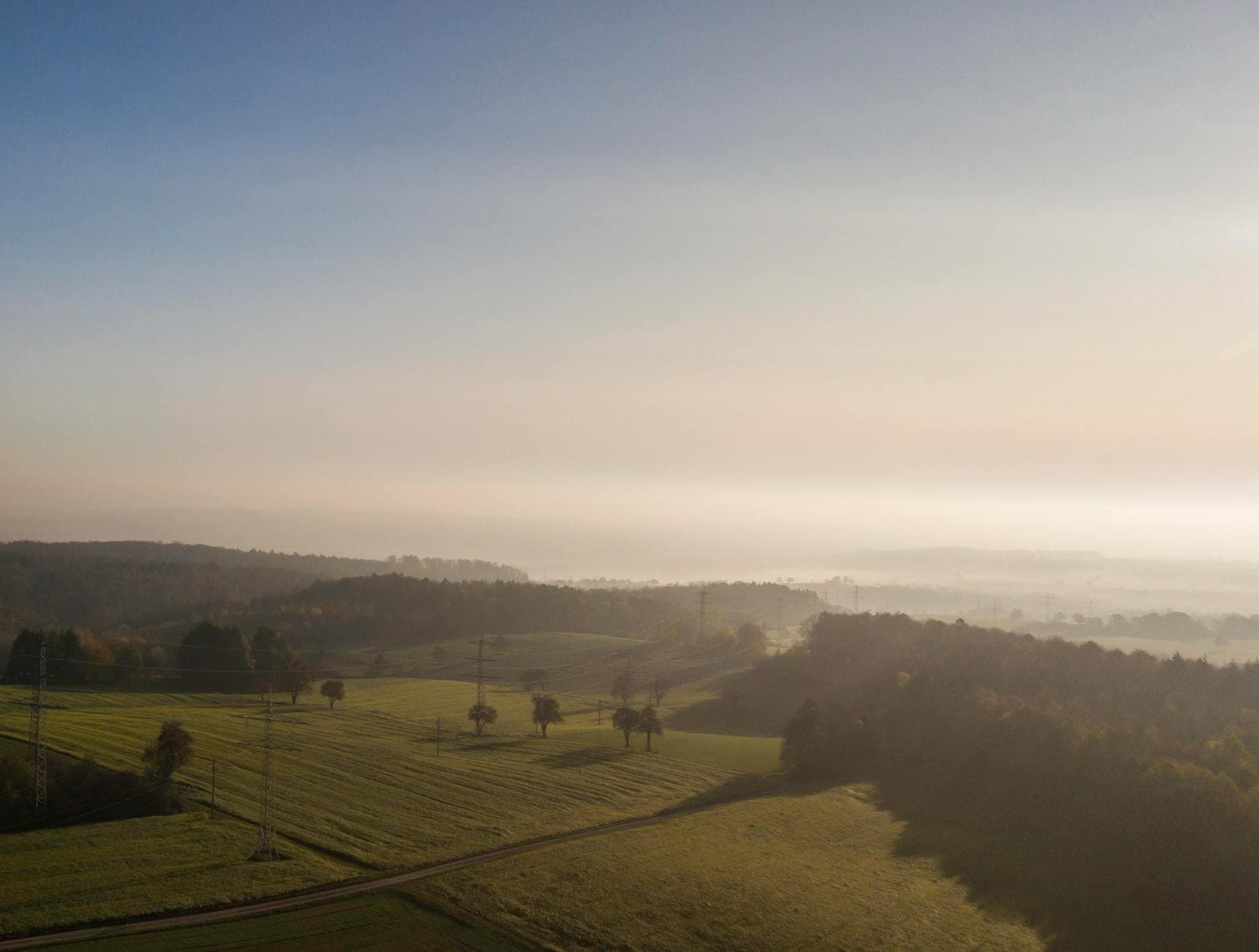 Morgendämmerung über hügeliger Landschaft mit Feldern, Bäumen, Strommasten und Nebelschwaden in der Ferne.