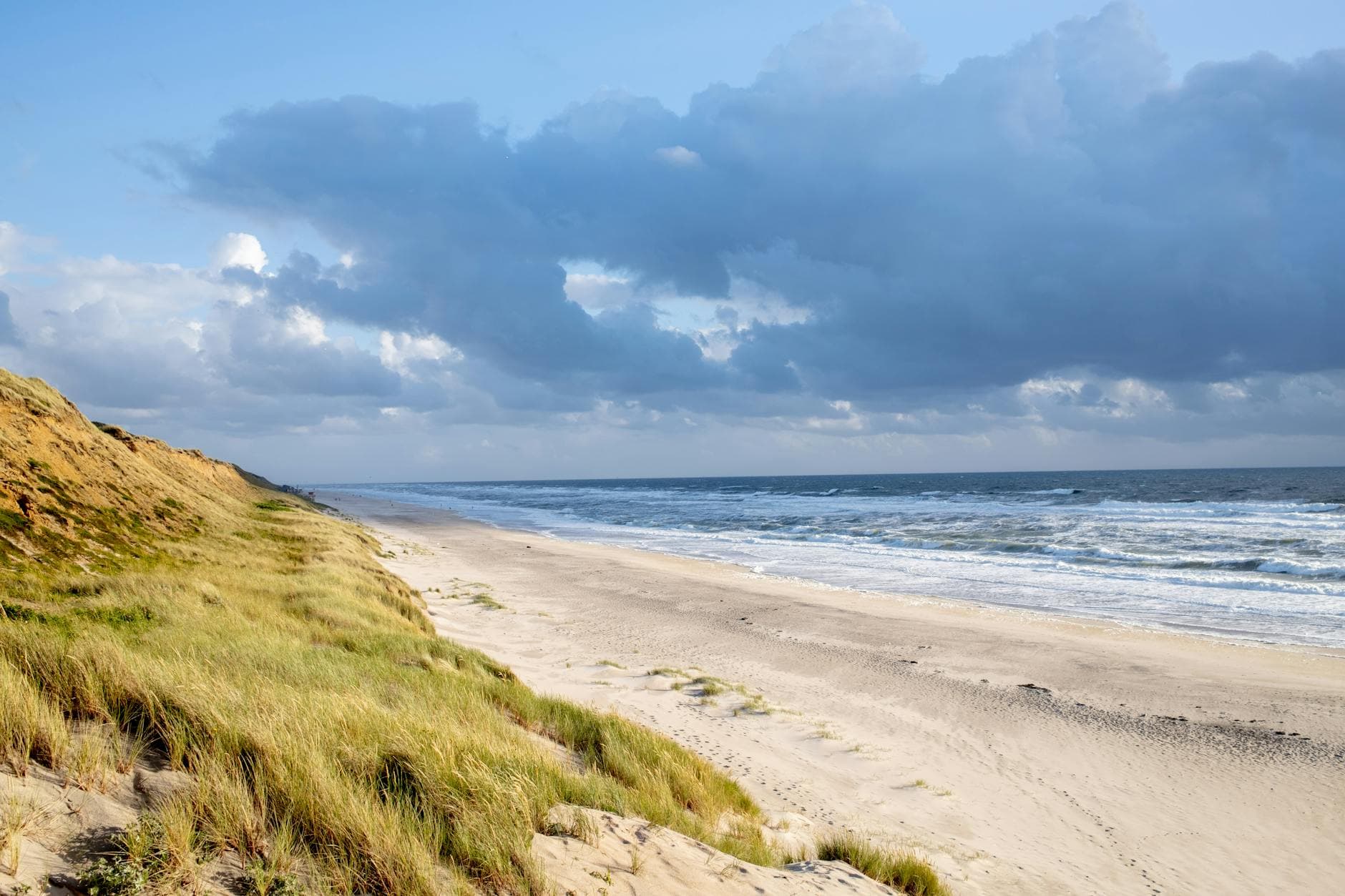 Strandlandschaft mit grasbewachsenen Sanddünen am Meer, dessen Wellen unter einem teils bewölkten Himmel brechen.