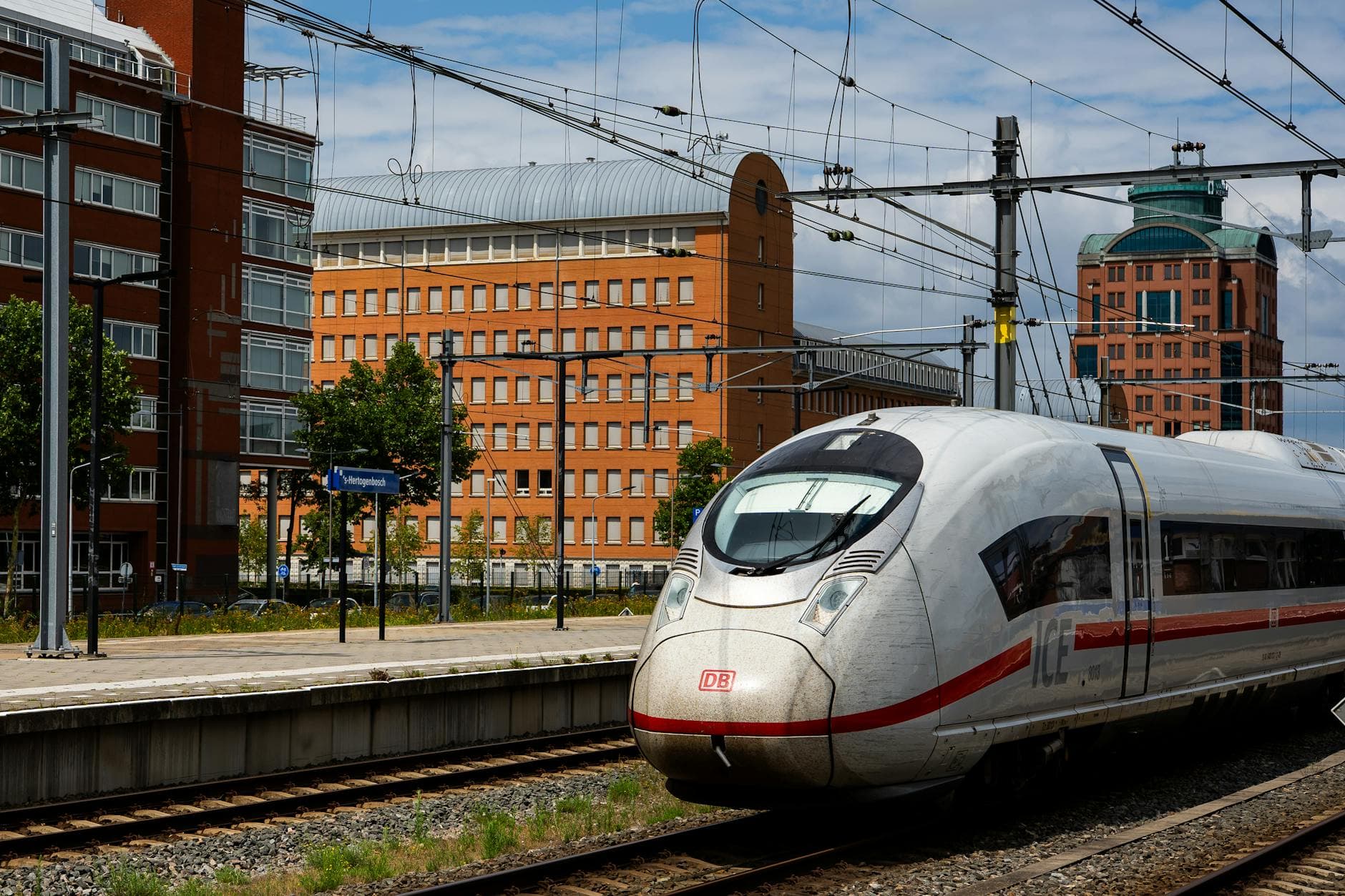 Weißer ICE-Zug mit DB-Logo am Bahnhofsgleis vor orangefarbenen Gebäuden, Oberleitungen und einem Schild 's-Hertogenbosch.