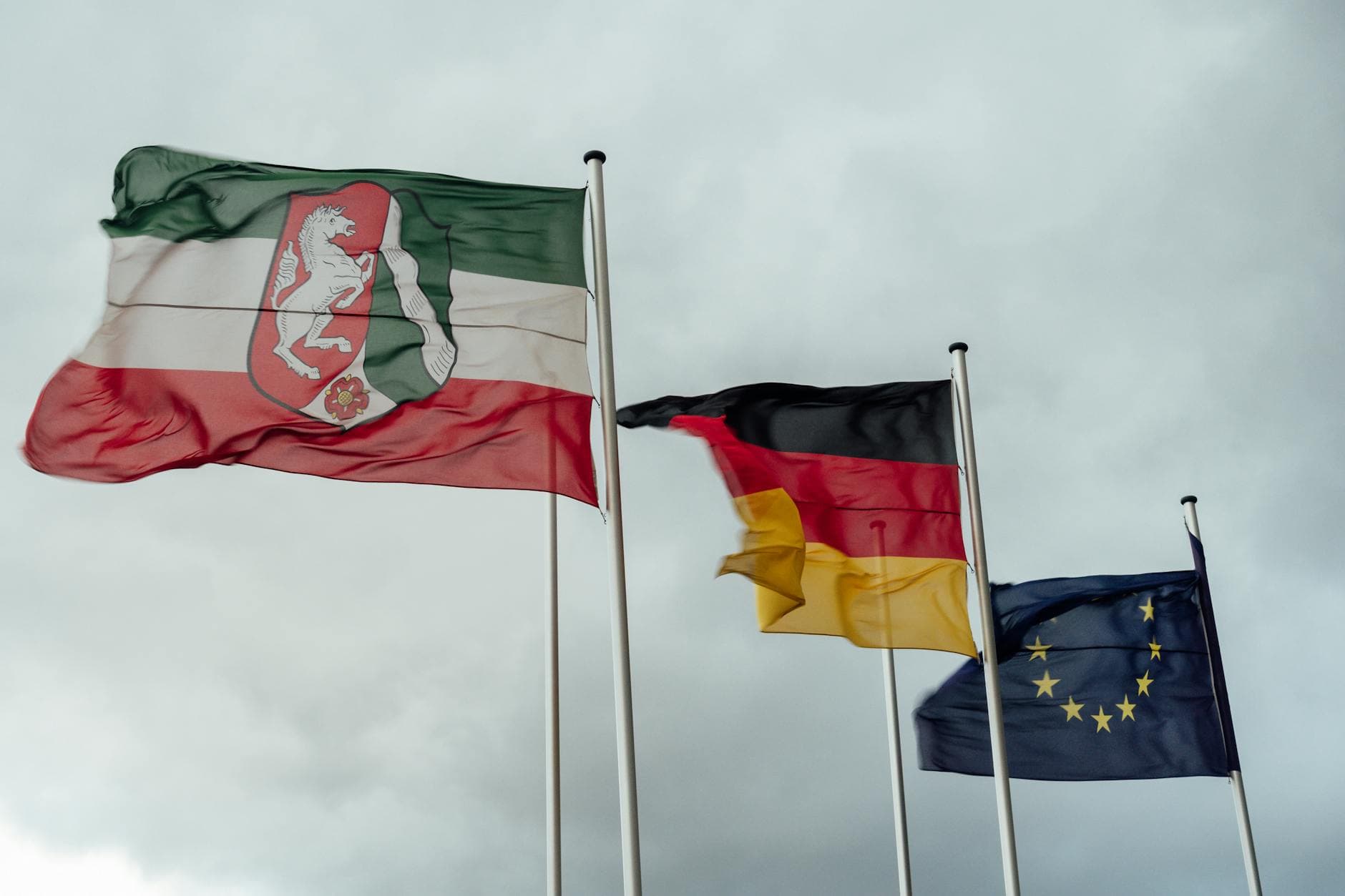 Wehende Flaggen von Nordrhein-Westfalen, Deutschland und der EU vor bewölktem Himmel.