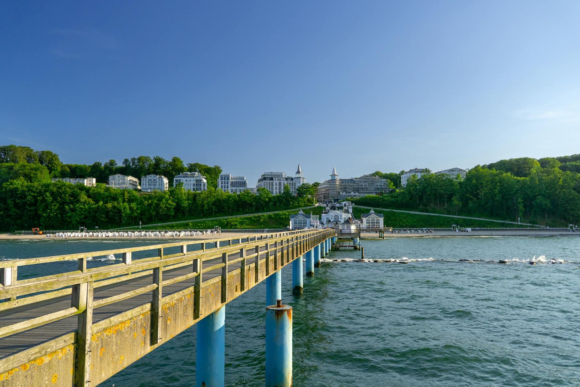 Hölzerne Seebrücke ragt ins blaue Meer, dahinter weißer Sandstrand mit Strandkörben und eleganten weißen Häusern auf bewaldetem Hügel unter blauem Himmel.